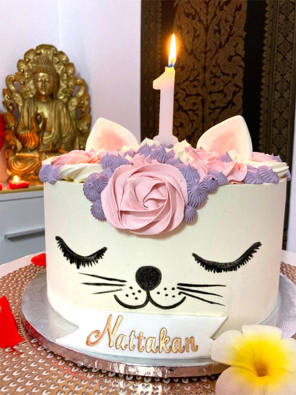 Una torta splendidamente decorata per assomigliare a un muso di gatto con occhi chiusi, naso nero, baffi e orecchie. La parte superiore della torta presenta decorazioni floreali rosa e viola e una singola candela con il numero uno, accesa in cima. Sullo sfondo, una statua dorata di Buddha circondata da candele. Alla base della torta c'è il nome Nattakan scritto su una targhetta bianca e oro.