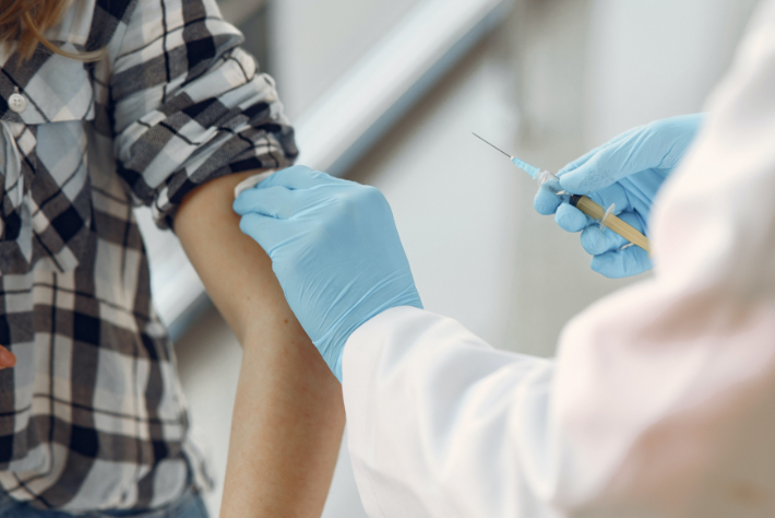 Een zorgverlener die blauwe handschoenen draagt, dient een injectie toe aan een persoon die een geruit overhemd draagt met opgerolde mouwen.