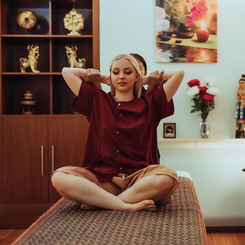 A woman receiving a traditional Thai massage while seated on a mat. Decor includes golden Thai statues on shelves in the background and a flower arrangement on a counter, creating a serene and relaxing atmosphere.