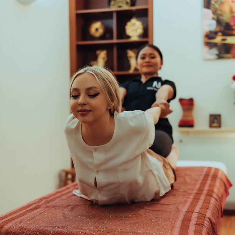 A female massage therapist is performing traditional Thai massage on a client lying on a massage bed at Nattakan Thai Massage salon. The client is smiling and appears relaxed as the therapist stretches her arms backward. The room is warmly lit with wooden shelves in the background displaying decorative items.