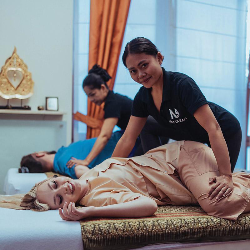 Two clients receiving Thai massages from therapists at Nattakan Thai Massage. One client in the foreground is a smiling woman lying on her side, being massaged by a therapist wearing a black Nattakan shirt. In the background, another therapist is massaging a clients back. The room is softly lit with orange curtains and traditional Thai decor.