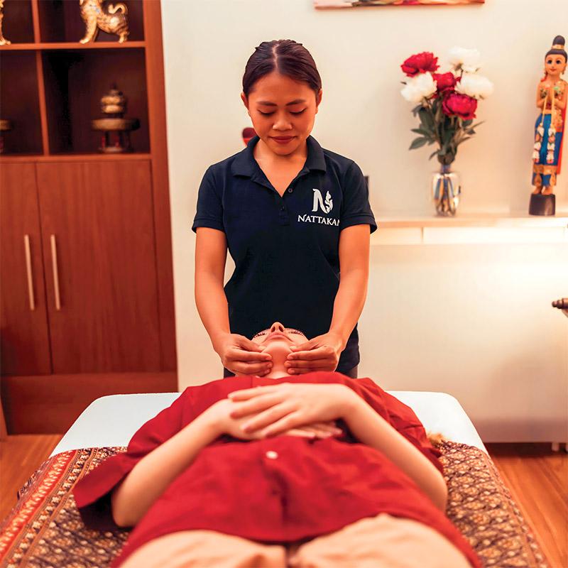 Một người phụ nữ mặc đồng phục Nattakan Thai Massage thực hiện massage mặt cho một khách hàng đang nằm trên bàn mát-xa. Căn phòng được trang trí bằng cách cắm hoa, tượng và kệ đựng đồ trang trí truyền thống.