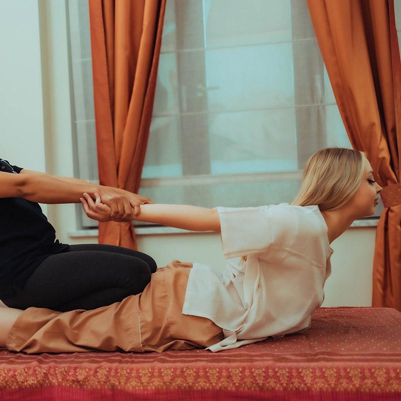 A woman receiving a traditional Thai massage where the therapist, positioned behind her, is gently pulling her arms backward while she lays on a massage mat. The room features warm, orange curtains and a peaceful ambiance.