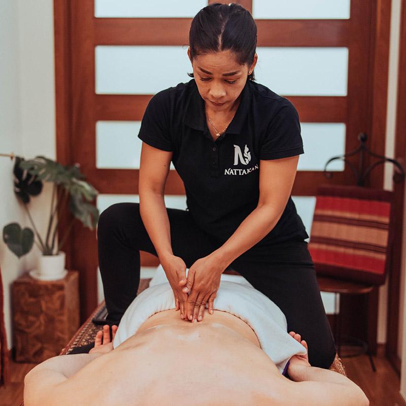 Nhân viên mát-xa người Thái mặc áo Nattakan Thai Massage màu đen thực hiện động tác mát-xa lưng kiểu Thái truyền thống cho khách hàng trong một căn phòng được trang trí đẹp mắt, thanh bình.