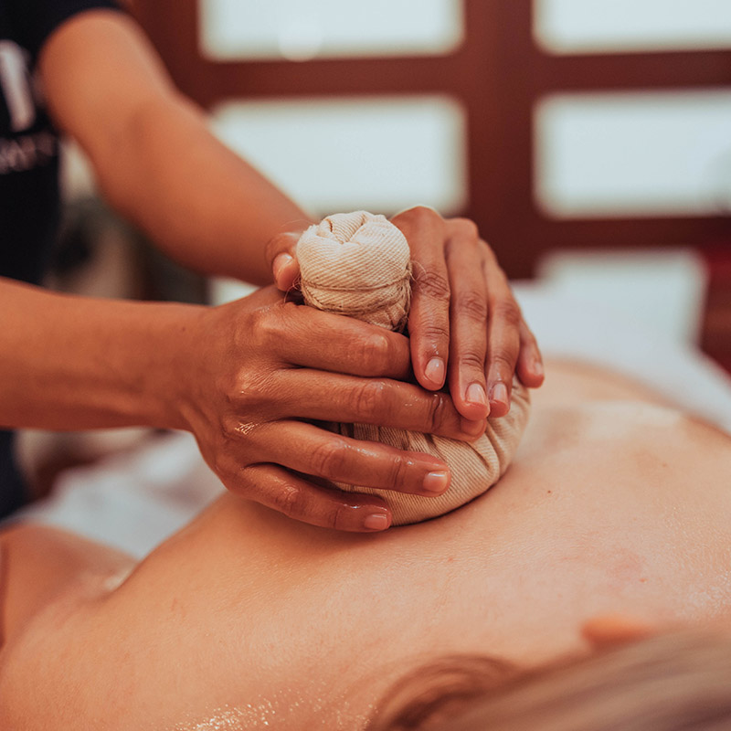 Foto close-up tangan terapis yang mengoleskan kompres herbal tradisional Thailand ke punggung seseorang saat memijat di salon Nattakan Thai Massage.