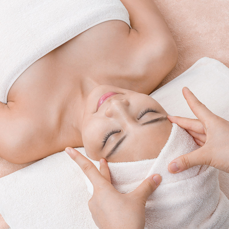 A person relaxing while receiving a facial massage, with their eyes closed and head wrapped in a towel at Nattakan Thai Massage salon.