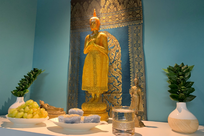 A tranquil shrine with a golden Buddha statue framed by an ornate blue and gold tapestry. The statue is adorned with a bright orange head decoration. On a white surface in front of the statue are two white vases holding green foliage, a plate with green grapes, and another plate with blue crystals. A smaller golden Buddha figure and a transparent candle holder with a gold accent are also present on the shrine.