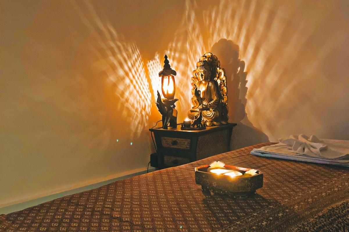 Una habitación serena en Nattakan Thai Massage, con una estatua de Buda suavemente iluminada y una linterna sobre una mesa de madera, con intrincados patrones de luz proyectados en la pared. En primer plano, una cama de masaje con toallas cuidadosamente dispuestas y una bandeja decorativa con velas encendidas y pétalos de flores.