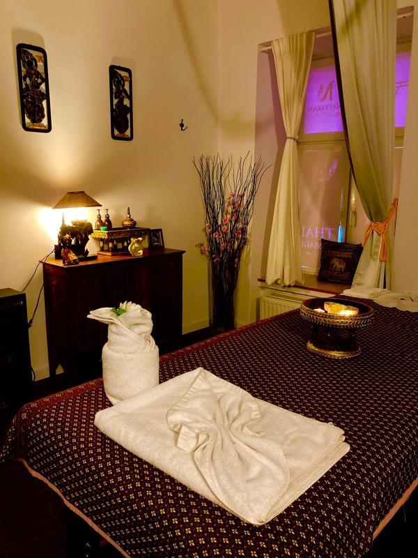 A tranquil Thai massage room featuring a neatly prepared massage bed covered with a patterned blanket, adorned with a folded white towel and a towel shaped like a swan. The room is softly lit by a small lamp on a wooden cabinet, which holds various decorative items and small statues. A window with partially open curtains allows a soft natural glow, enhanced by delicate decor like a tall vase with branches and flowers. The atmosphere is calm and inviting, perfect for a relaxing massage at Nattakan Thai Massage salon.