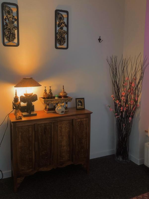 A tranquil corner of Nattakan Thai Massage salon featuring a wooden cabinet adorned with intricately carved designs, topped with traditional Thai decor items, a small lamp providing a warm glow, and a framed picture. Two vertical Thai art pieces hang on the wall above the cabinet. To the right of the cabinet, there is a tall, clear vase filled with decorative branches and pink blossoms, enhancing the serene and welcoming ambiance.