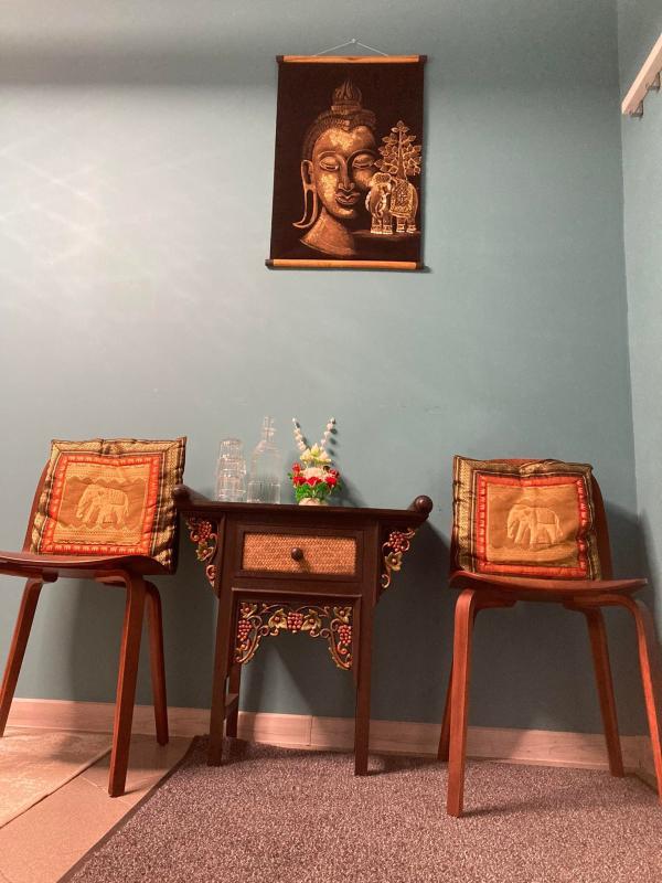 Cozy waiting area at Nattakan Thai Massage salon featuring two wooden chairs adorned with elephant-patterned cushions, a small ornate wooden table with a woven drawer, decorative glass bottles, and a floral arrangement. A tranquil Buddha tapestry hangs on the light blue wall above.