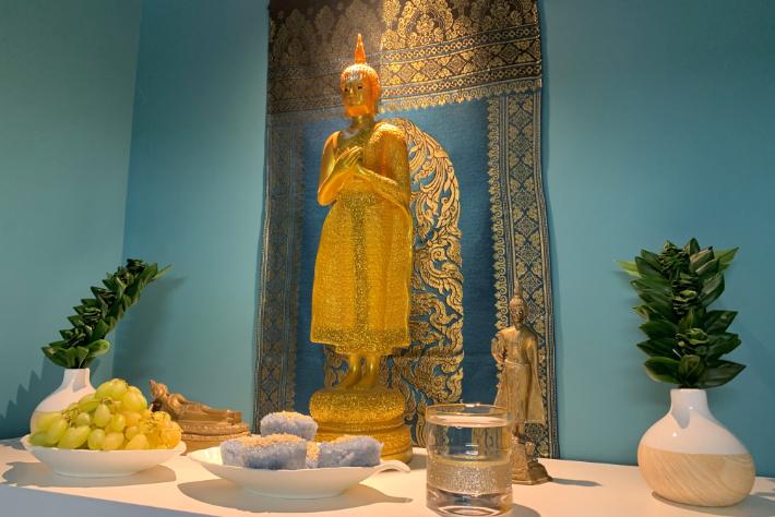 Alteropstilling med en gylden Buddhastatue med indviklede detaljer, placeret foran et udsmykket blåt og gyldent billedtæppe. Alteret er udsmykket med offergaver, bl.a. en skål med grønne druer, et fad med krystalformationer og en lille klar glasbeholder med et dekorativt mønster. To hvide vaser med grønne planter flankerer buddhastatuen og giver omgivelserne et strejf af naturlig elegance.