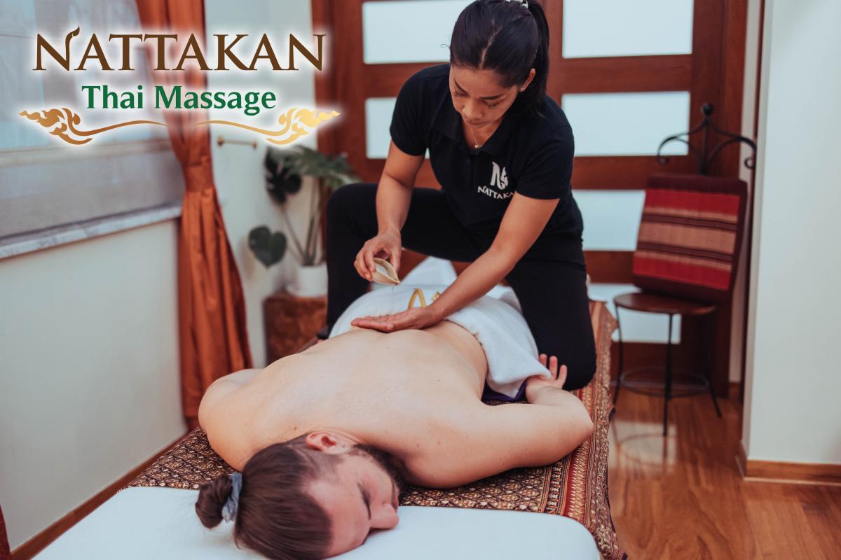 A massage therapist at Nattakan Thai Massage is giving a back massage using a warm herbal compress on a client lying face down on a massage table. The therapist is wearing a black uniform with the Nattakan logo. The room is decorated with warm wooden elements and a potted plant. The Nattakan Thai Massage logo is displayed in the upper left corner.