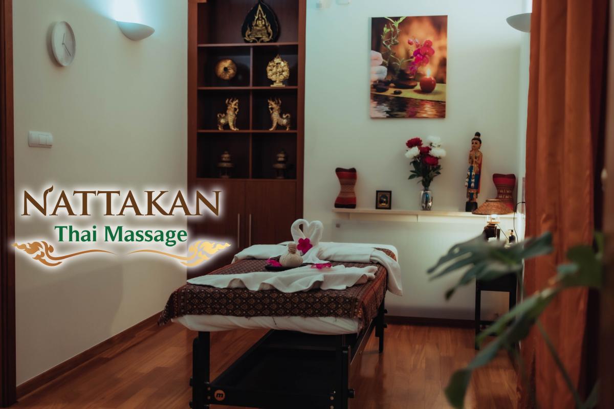 Privat massasjerom med massasjebord med dekorative brettede håndklær og blomsterblader, trehyller med tradisjonelle thailandske ornamenter, en veggklokke og Nattakan Thai Massages logo i forgrunnen.