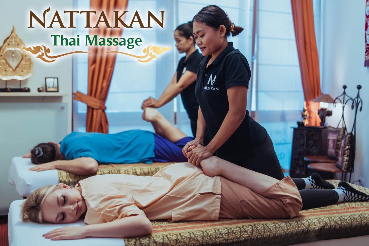 Two individuals receiving a Thai massage at Nattakan Thai Massage salon. A therapist in a black shirt with the Nattakan logo provides a massage, stretching one client’s legs. The calm and serene massage room features warm lighting and traditional decor.