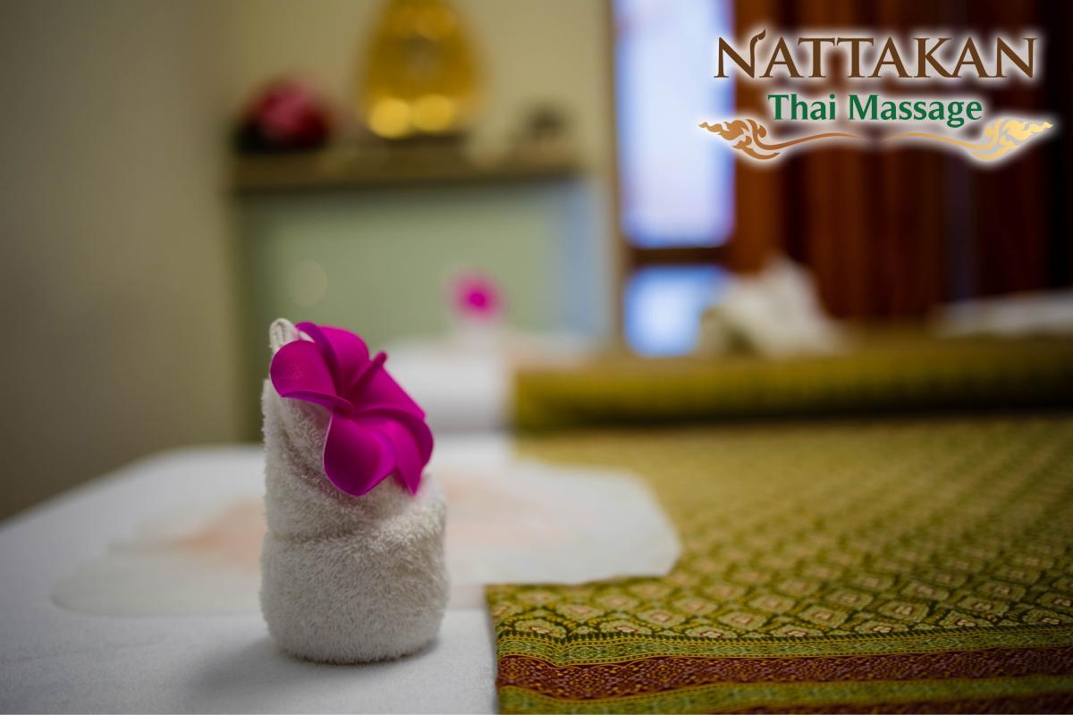 Close-up of a neatly folded white towel with a bright pink flower on top, set on a massage table covered with a patterned green and gold traditional cloth at Nattakan Thai Massage salon. The background is softly blurred, focusing attention on the towel and flower.