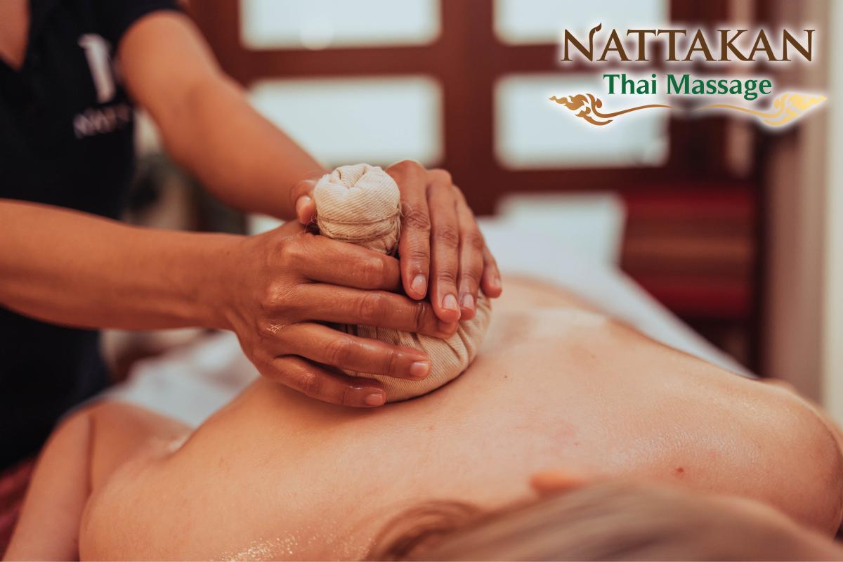 Un terapeuta esegue un massaggio tailandese con impacco alle erbe sulla schiena di una persona al Nattakan Thai Massage. Il terapeuta utilizza un impacco alle erbe avvolto in un panno, applicandolo delicatamente sulla schiena oliata del cliente. Il logo del Nattakan Thai Massage è visibile nell'angolo in alto a destra dell'immagine.