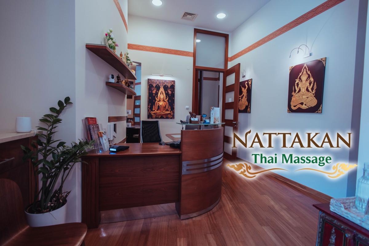 Khu vực lễ tân của Nattakan Thai Massage salon có bàn lễ tân bằng gỗ hiện đại, cây trang trí, tạp chí và các tác phẩm nghệ thuật truyền thống của Thái Lan trên tường. Logo Nattakan Thai Massage hiển thị trong hình ảnh. Không gian được chiếu sáng ấm áp và có bầu không khí thân thiện.