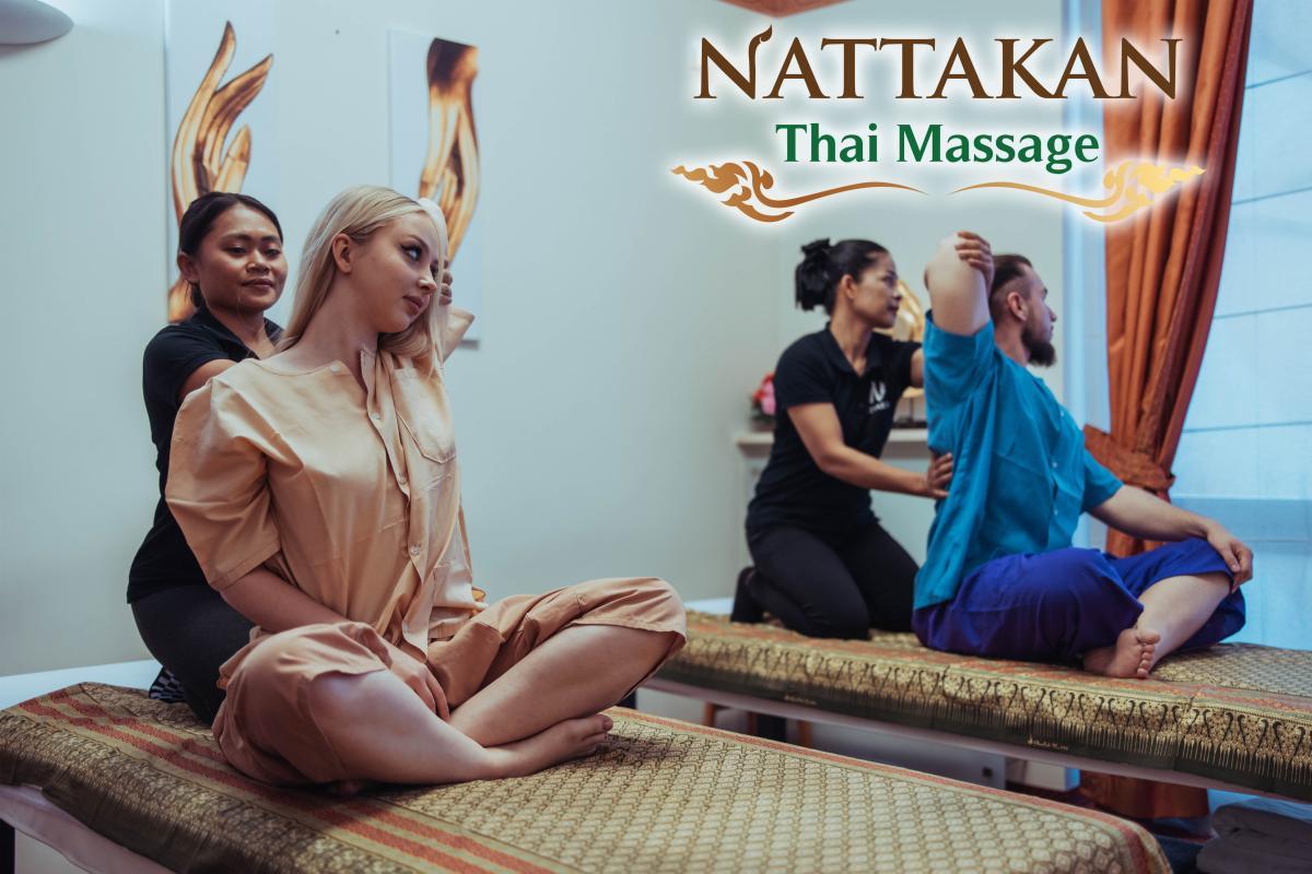 Zwei Therapeuten geben einem Mann und einer Frau, die auf Massagebetten in einem ruhigen Raum sitzen, Thai-Massagen. Das Logo von Nattakan Thai Massage befindet sich in der oberen rechten Ecke des Bildes.