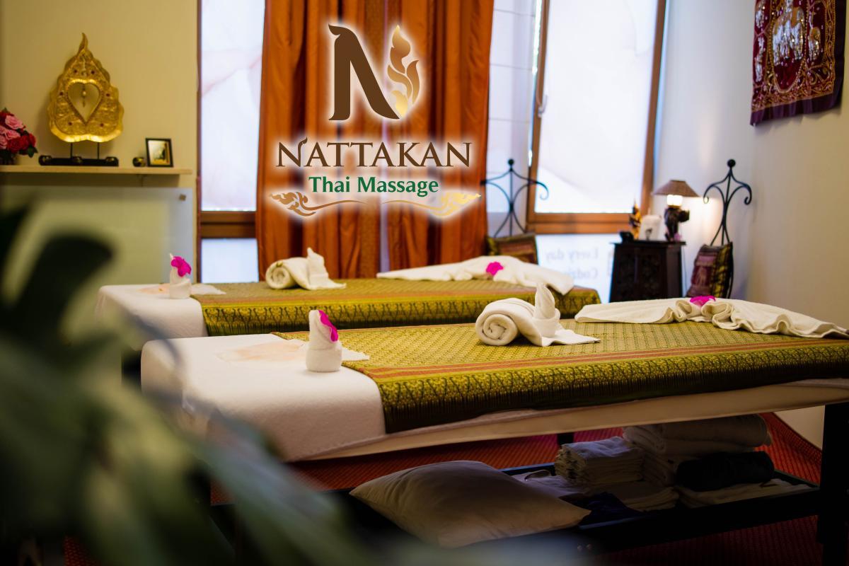 Quang cảnh bên trong phòng mát-xa yên tĩnh tại Nattakan Thai Massage, có hai bàn mát-xa được chuẩn bị gọn gàng với những chiếc khăn cuộn và điểm nhấn bằng hoa. Căn phòng được chiếu sáng ấm áp với ánh sáng dịu nhẹ, lối trang trí truyền thống của Thái Lan và bầu không khí thanh bình. Logo Nattakan Thai Massage được hiển thị nổi bật ở giữa hình ảnh.