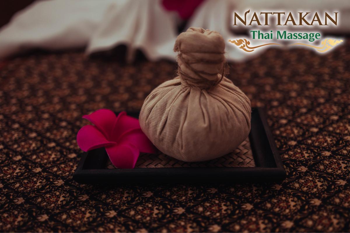 A herbal compress ball and a red flower placed on a woven tray, set on a patterned fabric surface. The logo of Nattakan Thai Massage is visible in the upper right corner.