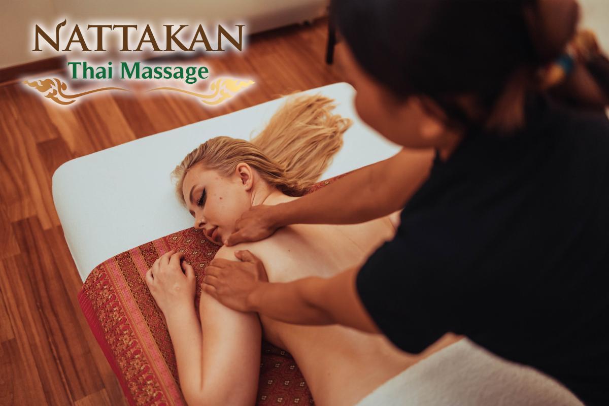 Une personne recevant un massage thaïlandais sur le dos à Nattakan Thai Massage. Le client est allongé sur le ventre sur une table de massage, recouverte d'un tissu traditionnel à motifs. Les mains de la masseuse exercent une pression sur les épaules du client. Le logo de Nattakan Thai Massage est visible dans le coin supérieur gauche de l'image.