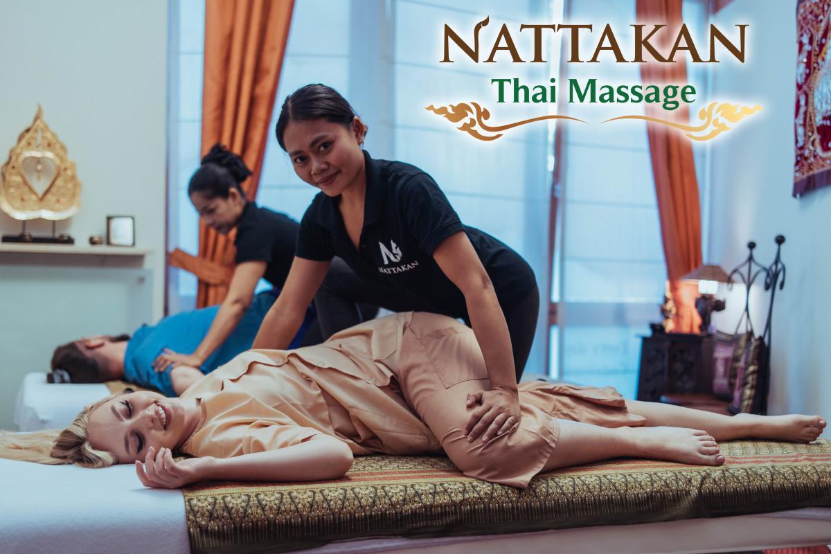 Two people receiving Thai massages at Nattakan Thai Massage salon. The foreground shows a woman in beige clothing smiling while receiving a massage on a traditional Thai mat, given by a therapist in a black shirt with the Nattakan logo. In the background, another woman in similar attire is providing a massage to a man lying on a table. The room is softly lit with natural light from a window adorned with orange curtains, and there are decorative elements on the walls and a small shelf.