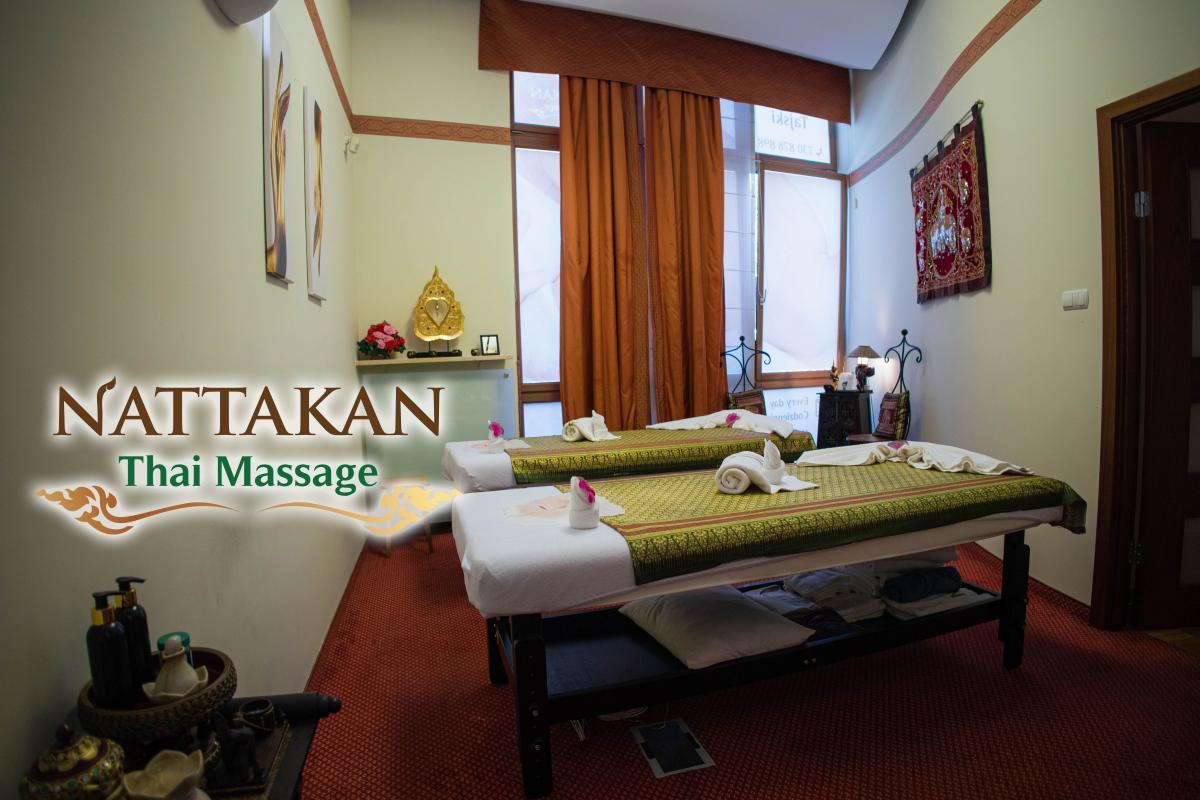 Interior of Nattakan Thai Massage salon showcasing a serene room with two massage beds decorated with green and gold linens, neatly folded towels, and pink flowers. The room features soft lighting, warm-toned curtains, traditional Thai decor, and a shelf with a golden decorative piece and flowers. The logo of Nattakan Thai Massage is displayed in the image.