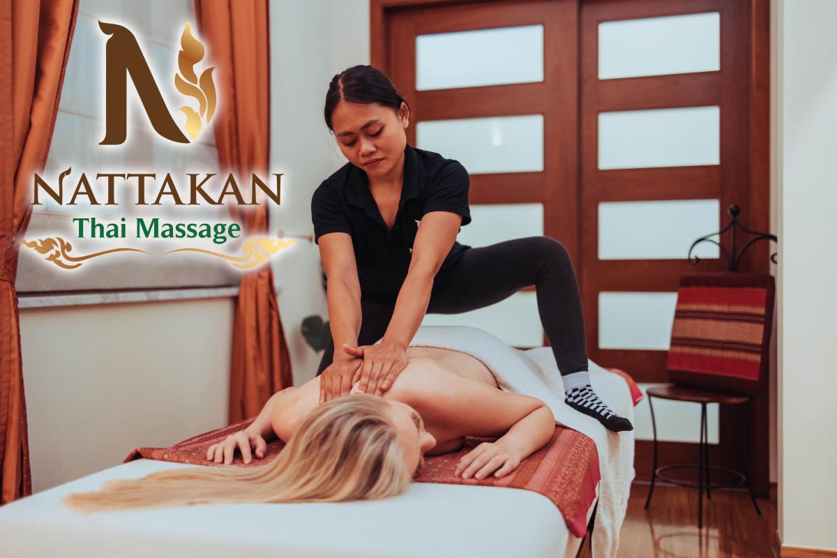 Massage therapist performing a traditional Thai massage on a client lying on a massage table at Nattakan Thai Massage salon. The therapist is pressing the clients back with their hands, while the client is relaxed and lying face-down. The Nattakan Thai Massage logo is displayed on the left side of the image.