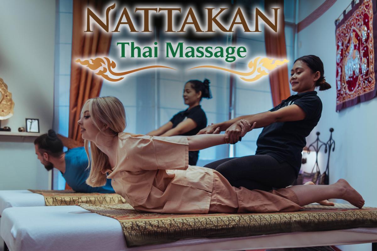 Two clients, a man and a woman, receiving traditional Thai massages at Nattakan Thai Massage salon. The therapists perform a stretching technique while the clients lie on massage mats in a serene and beautifully decorated room.