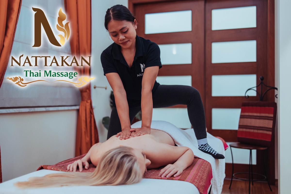 Terapeutka wykonująca masaż tajski w salonie Nattakan Thai Massage. Terapeutka używa techniki nacisku dłońmi na plecach klientki leżącej na stole do masażu. W tle widoczne są czerwone zasłony i drewniane drzwi. Po lewej stronie nałożone jest logo Nattakan Thai Massage.