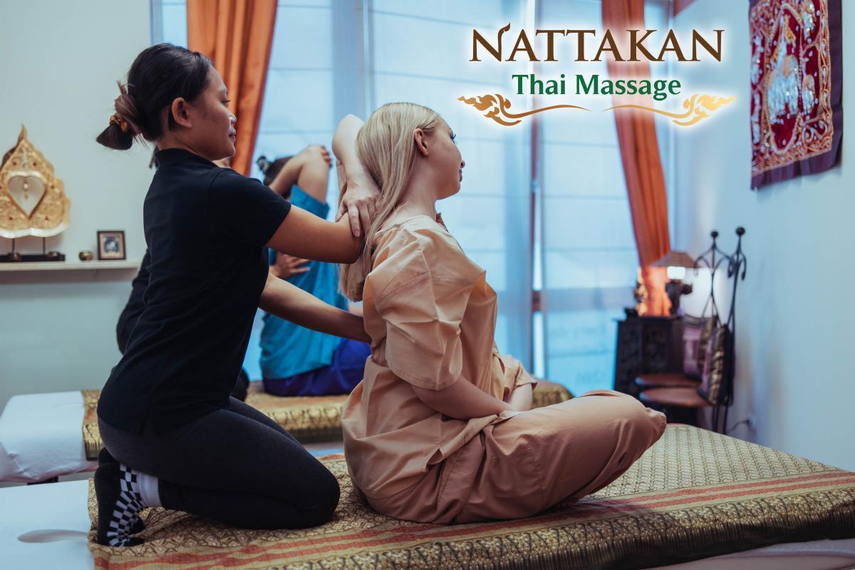 A professional therapist performing a traditional Thai massage on a client dressed in comfortable attire at Nattakan Thai Massage salon. The room is softly lit, decorated with Thai-themed decor, including ornate wall art and a small shelf with cultural ornaments. The client is seated on a padded mat while the therapist is kneeling behind, stretching the clients shoulders. The Nattakan Thai Massage logo is visible in the upper right corner of the image.