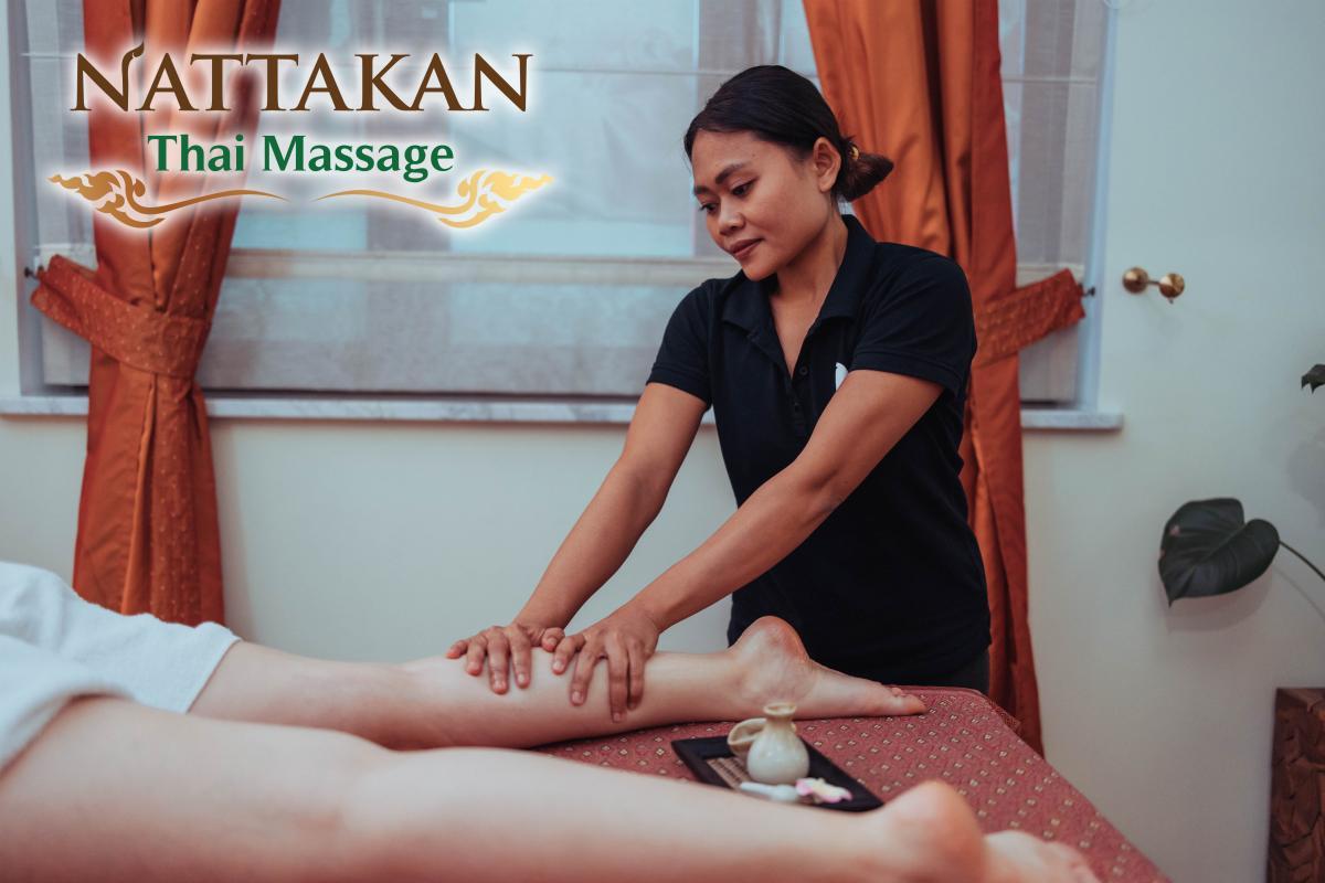 Un terapeuta pratica un massaggio alle gambe a un cliente in una stanza tranquilla del salone Nattakan Thai Massage, con il logo del salone ben visibile nell'angolo in alto a sinistra. L'ambiente è caratterizzato da tende dai toni caldi e luci soffuse.