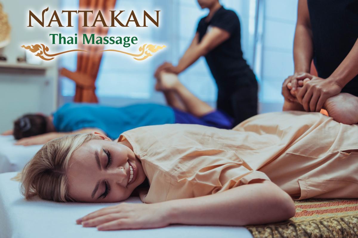 Nattakan Thai Massage'da masaj yaptıran bir kişi masaj masasına yüzüstü uzanmış, gülümsüyor ve rahatlamış görünüyor. Arka planda başka bir kişi de ayrı bir masada masaj yaptırıyor. Nattakan Thai Masajı logosu görüntünün sol üst köşesinde yer almaktadır.