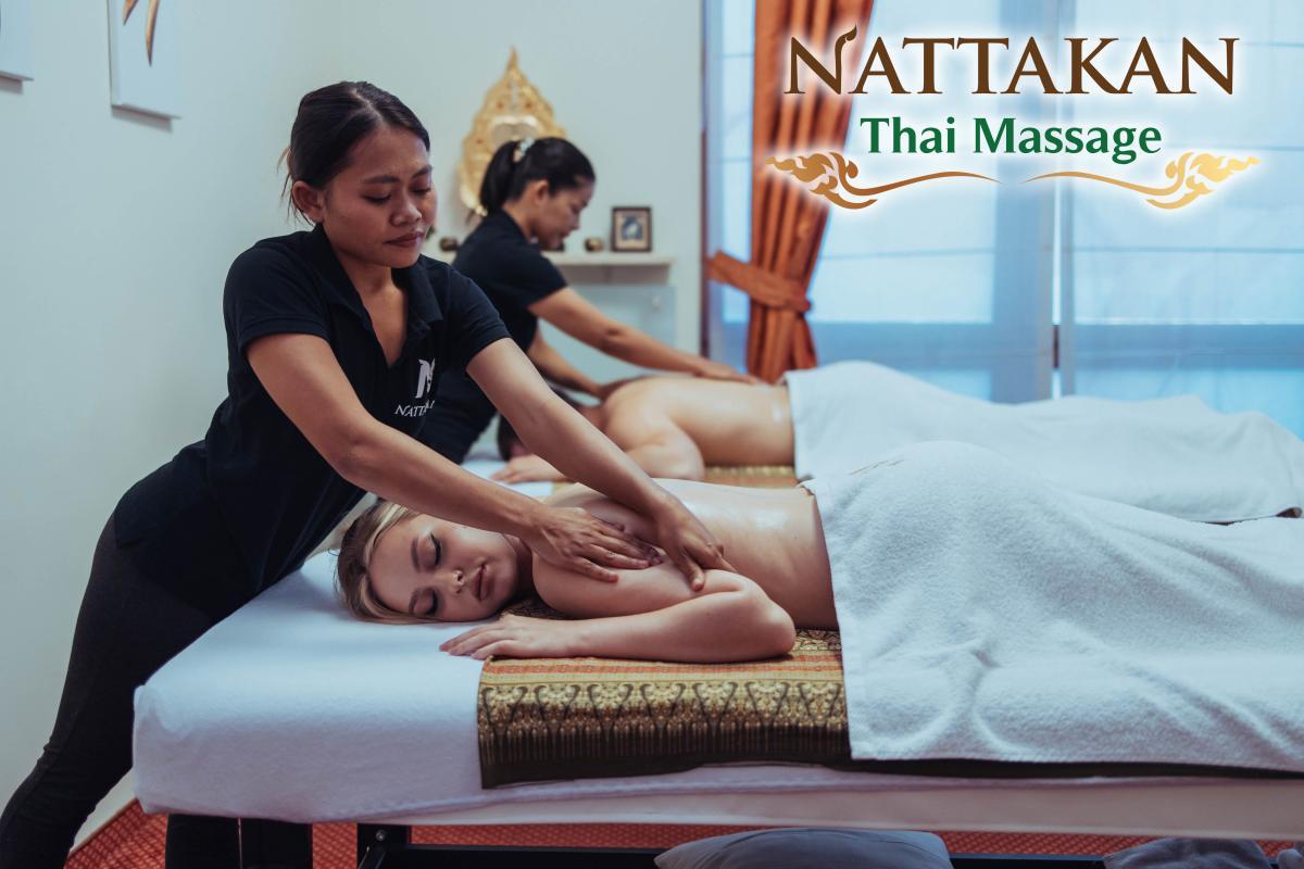 Massage therapists providing Thai massages to clients at Nattakan Thai Massage salon. Clients are lying on massage tables covered with white towels in a serene room with soft lighting and traditional Thai decor. The Nattakan Thai Massage logo is displayed in the top right corner.