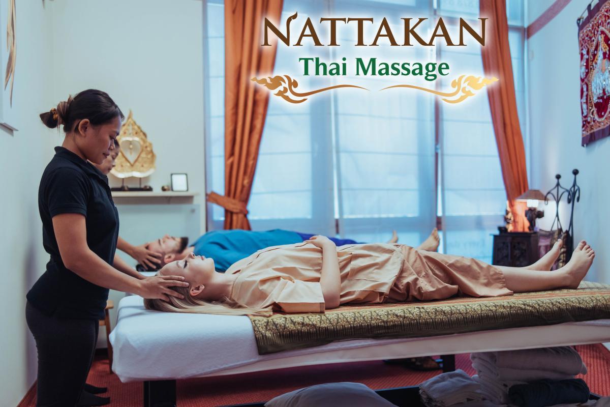 Terapis pijat melakukan pijatan di kepala klien yang sedang bersantai di tempat tidur pijat di Nattakan Thai Massage. Klien lain sedang menerima pijatan di latar belakang. Ruangan ini memiliki pencahayaan yang hangat dan dekorasi yang terinspirasi dari Thailand.