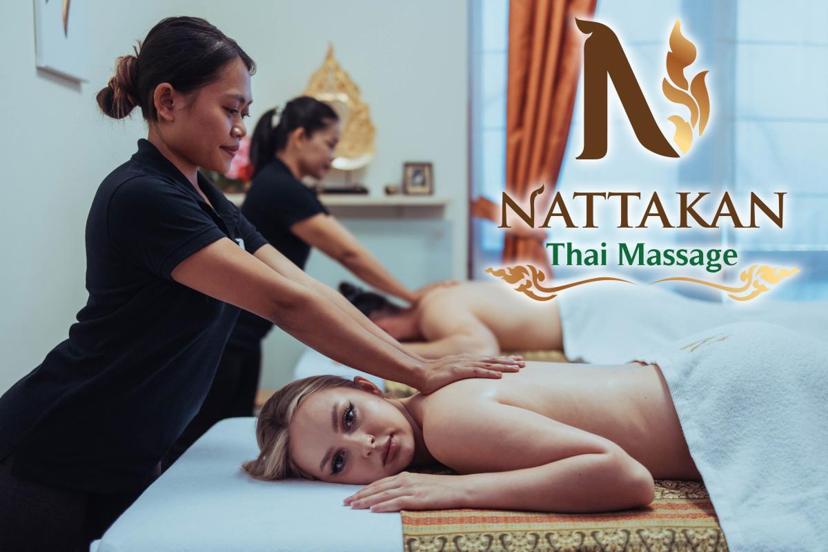 Kaksi naista saa hierontaa Nattakan Thai Massage -salongissa kahden ammattitaitoisen terapeutin antamana. Huone on rauhallinen ja sisustettu lämpimillä väreillä, jotka lisäävät rentouttavaa tunnelmaa. Nattakan Thai Massage -logo näkyy näkyvästi kuvan oikealla puolella.