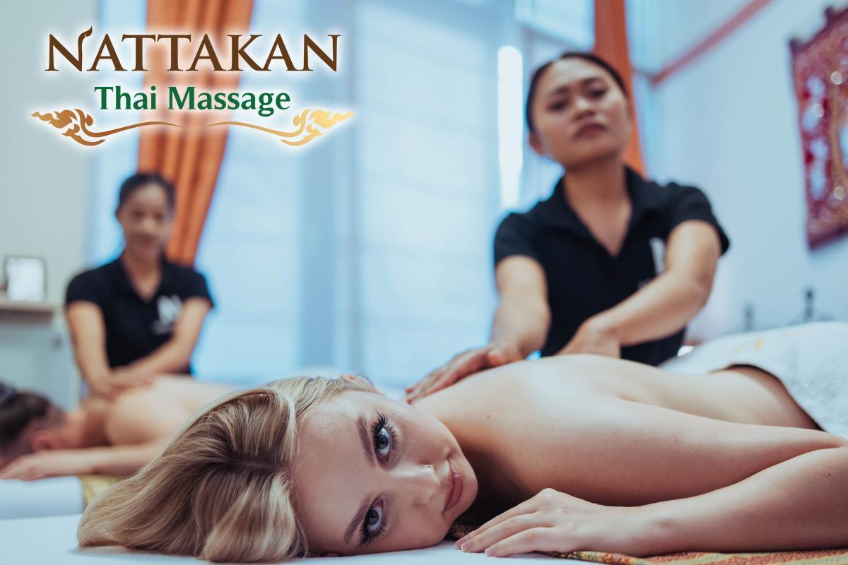 Two women receiving back massages from therapists in a serene room with soft lighting and warm-toned decor. The Nattakan Thai Massage logo is displayed in the top left corner. The foreground woman, lying face down on a massage table, looks relaxed and content.