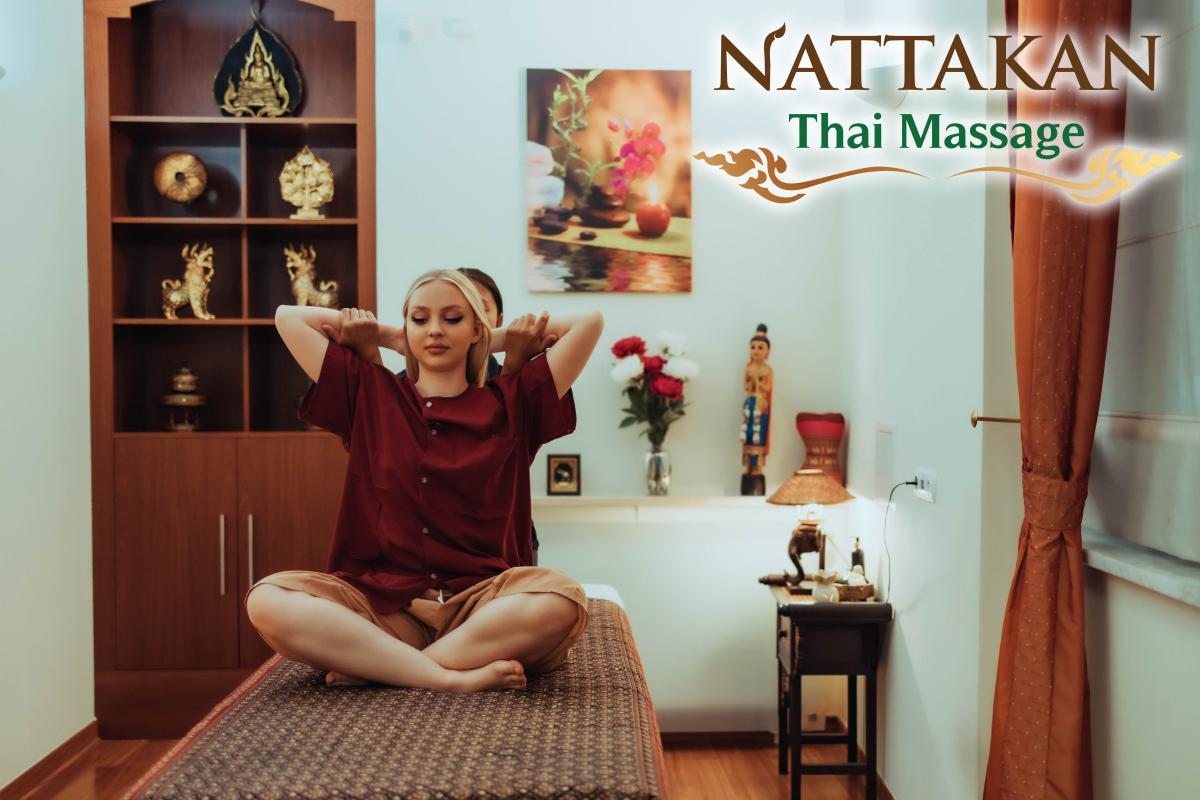 A woman sits cross-legged on a massage table, with a massage therapist providing a Thai massage by stretching her arms behind her head. The room is decorated with golden Buddha statues, traditional Thai decorations, and a shelf with ornate items. The Nattakan Thai Massage logo is displayed on the upper right corner.