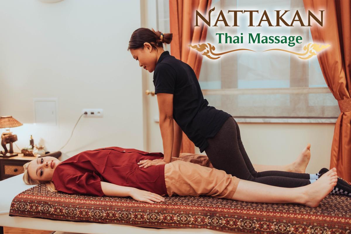 Một học viên massage Thái trong bộ trang phục màu đen đang thực hiện massage Thái truyền thống cho một khách hàng đang nằm trên bàn massage được trang trí. Khách hàng mặc áo đỏ và quần màu be, trông có vẻ thoải mái trong khung cảnh phòng spa yên tĩnh với ánh sáng dịu nhẹ và lối trang trí nhẹ nhàng. Logo của Nattakan Thai Massage hiển thị ở góc trên bên phải.