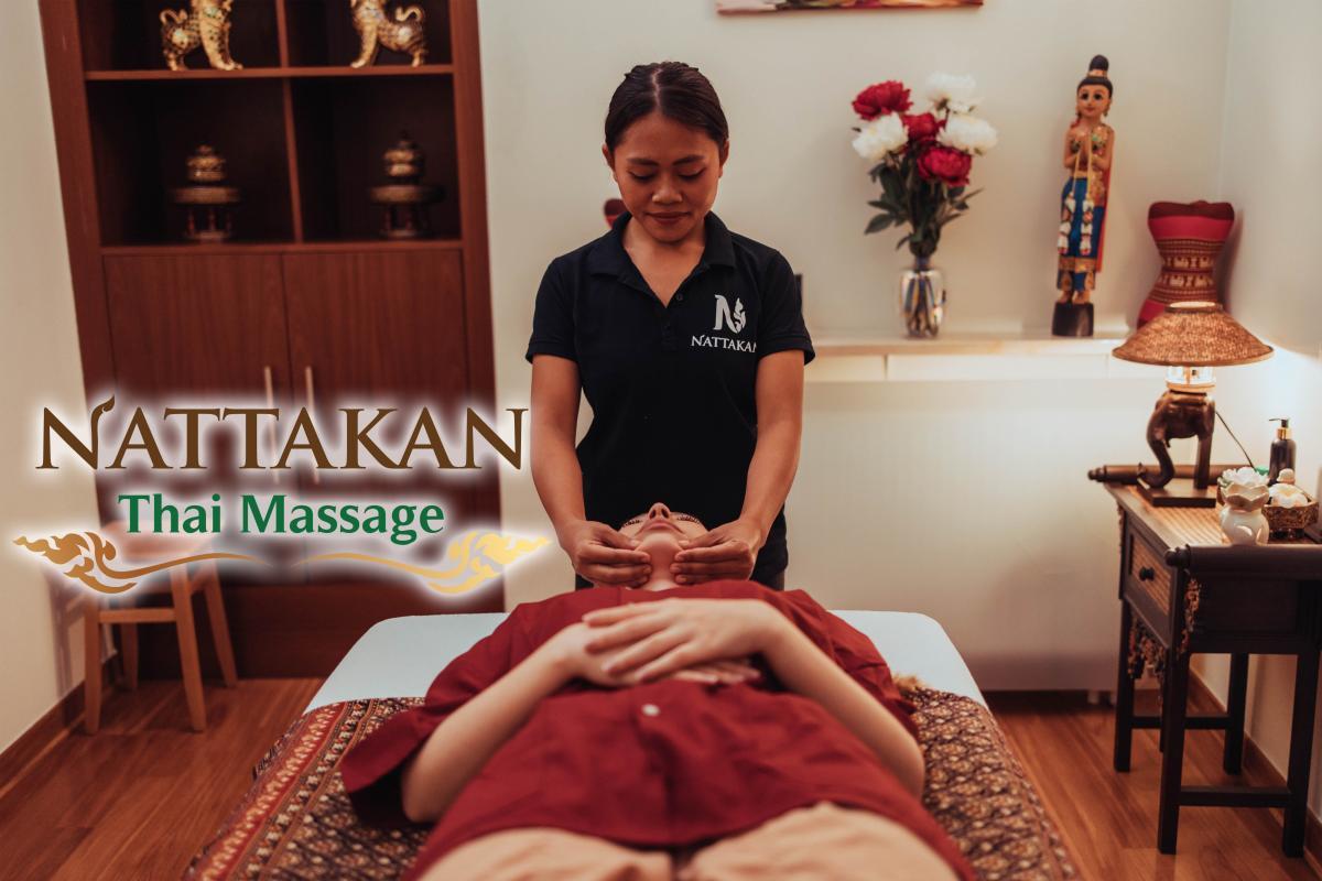 En massageterapeut på Nattakan Thai Massage utför en ansiktsmassage på en kund som ligger på en massagebänk. Rummet är varmt upplyst och dekorerat med thailändska kulturelement, bland annat en staty, en vas med blommor och en bordslampa. Terapeuten bär en svart uniform med Nattakans logotyp. Nattakan Thai Massages logotyp visas tydligt på bilden.