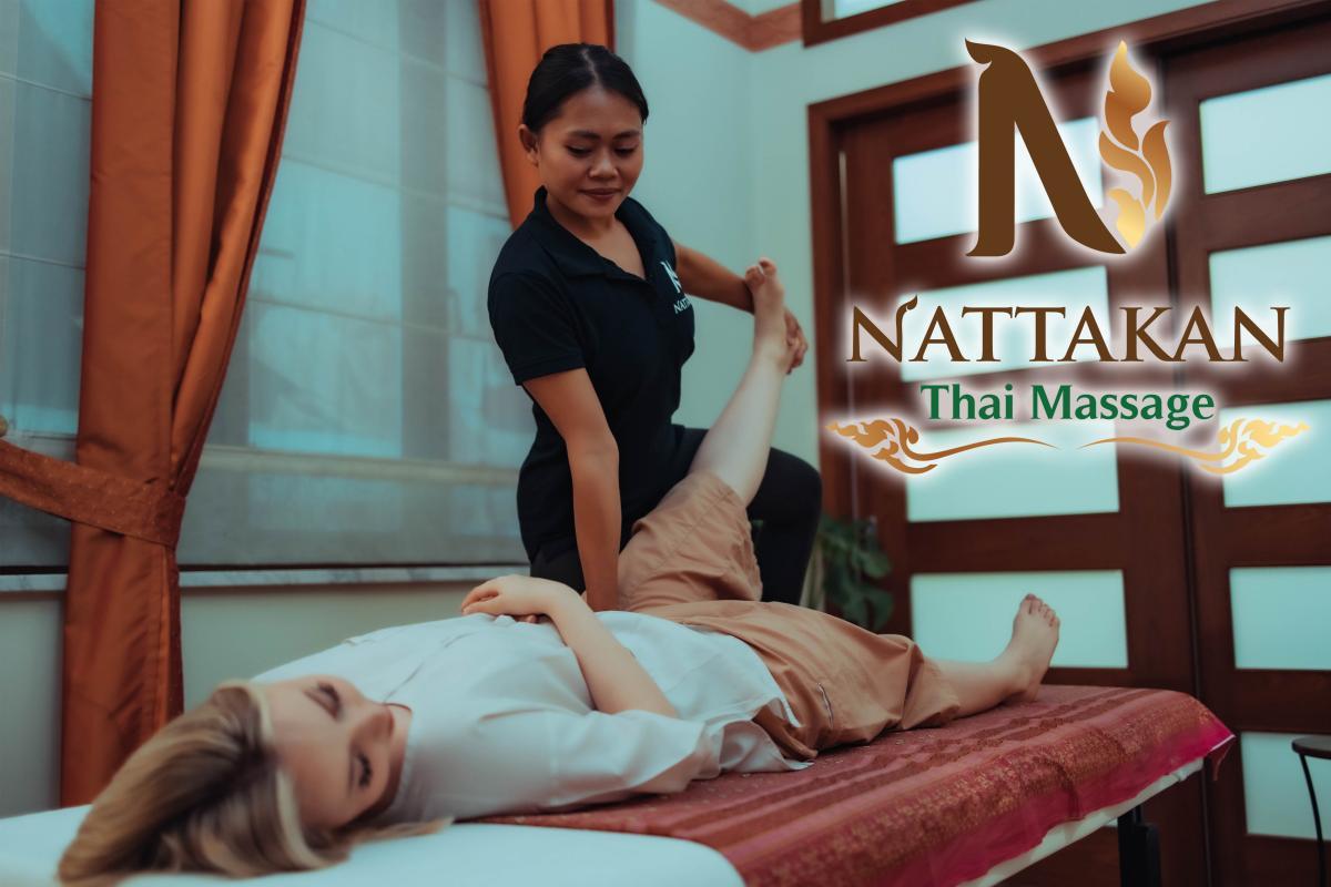 Một nhà trị liệu mát-xa tại Nattakan Thai Massage đang thực hiện liệu pháp mát-xa kiểu Thái truyền thống cho một khách hàng đang nằm trên giường mát-xa trong một căn phòng thanh bình có rèm màu cam. Logo Nattakan Thai Massage hiển thị trong hình ảnh.