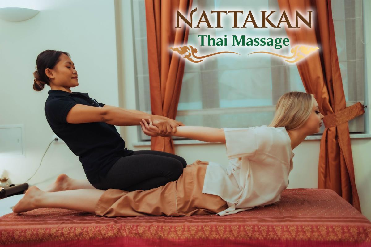 A massage therapist at Nattakan Thai Massage salon performs a traditional Thai massage on a client, who is lying on a massage bed. The therapist is stretching the clients arms and back while the client appears to be relaxed. The setting of the room is calm, with warm colored curtains and soft lighting. The Nattakan Thai Massage logo is visible at the top right corner of the image.