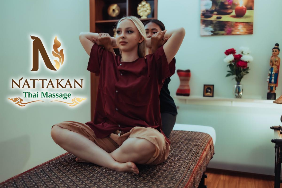 Client receiving a traditional Thai massage at Nattakan Thai Massage, performed in a serene room with soft lighting, decorative flowers, and cultural statues. The client is seated on a massage table with their arms being gently stretched by the therapist. The Nattakan Thai Massage logo is prominently displayed on the left side of the image.