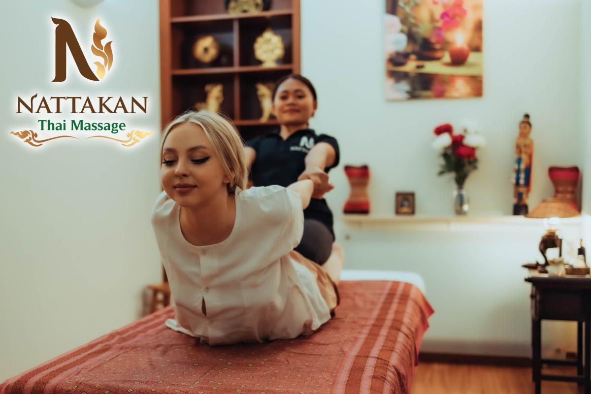 Kobieta otrzymująca masaż tajski w salonie Nattakan Thai Massage. Masażystka delikatnie naciąga ramiona klientki, która leży na stole do masażu. Po lewej stronie widoczne jest logo Nattakan. W tle znajduje się regał z dekoracjami oraz stół z kwiatami i figurkami.