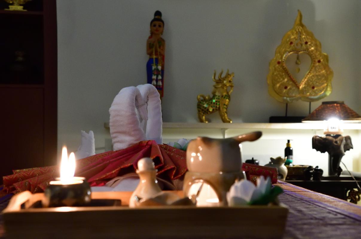 A cozy Thai massage room featuring a candle-lit ambiance. Two swan-shaped towel arrangements rest on a massage table adorned with a red cloth. Traditional Thai ornaments, including a golden statue and a Buddha figure, decorate the background. A tray of massage oils and ceramic containers is placed in the foreground. The setting is warm and inviting, reflective of a serene atmosphere.