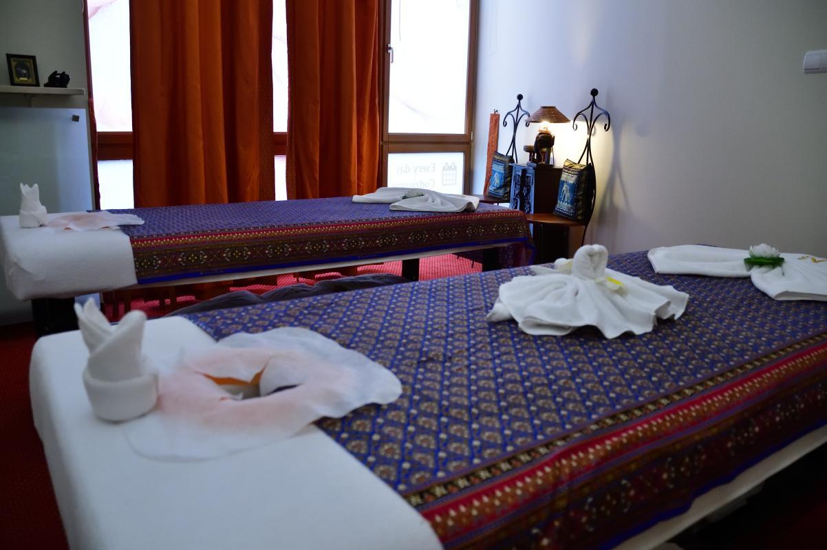 Una sala de masajes serena y bien decorada en Nattakan Thai Massage, con dos camas de masajes con fundas azules y rojas estampadas. En cada cama hay toallas cuidadosamente dobladas con formas de animales y flores. La habitación está suavemente iluminada con luz cálida y las ventanas están cubiertas con cortinas naranjas. Al fondo se ve una estantería con artículos de decoración y almohadas, que contribuyen a crear un ambiente relajante.