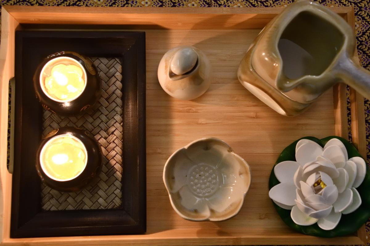 A wooden tray containing two lit candles in black holders, a ceramic pot with a lid, a ceramic jug, a flower-shaped ceramic bowl, and a white artificial lotus flower with green leaves. The arrangement exudes a calming and serene ambiance, perfect for a spa or massage setting.