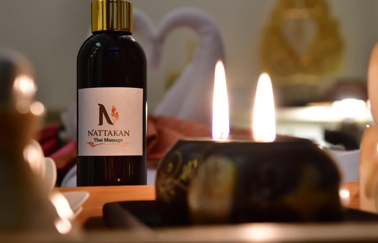 A bottle of massage oil labeled Nattakan Thai Massage is placed on a wooden surface. In the background, two softly lit candles add a serene and calming ambiance. The setting suggests a relaxing and peaceful environment ideal for a massage session.