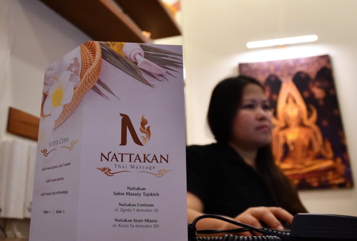 Primer plano de un folleto de Nattakan Thai Massage. El folleto, situado en primer plano, muestra de forma destacada el logotipo de Nattakan Thai Massage y la información de contacto de sus locales. En el fondo, una persona trabaja en un mostrador de una sala decorada con una imagen de Buda.